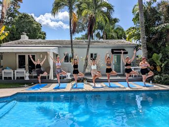 Group of eight women doing outdoor yoga tree pose on blue mats beside a sunny tropical backyard pool with palm trees and a white cottage under a clear blue sky.