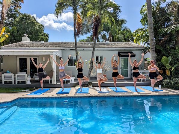 Group of eight women doing outdoor yoga tree pose on blue mats beside a sunny tropical backyard pool with palm trees and a white cottage under a clear blue sky.