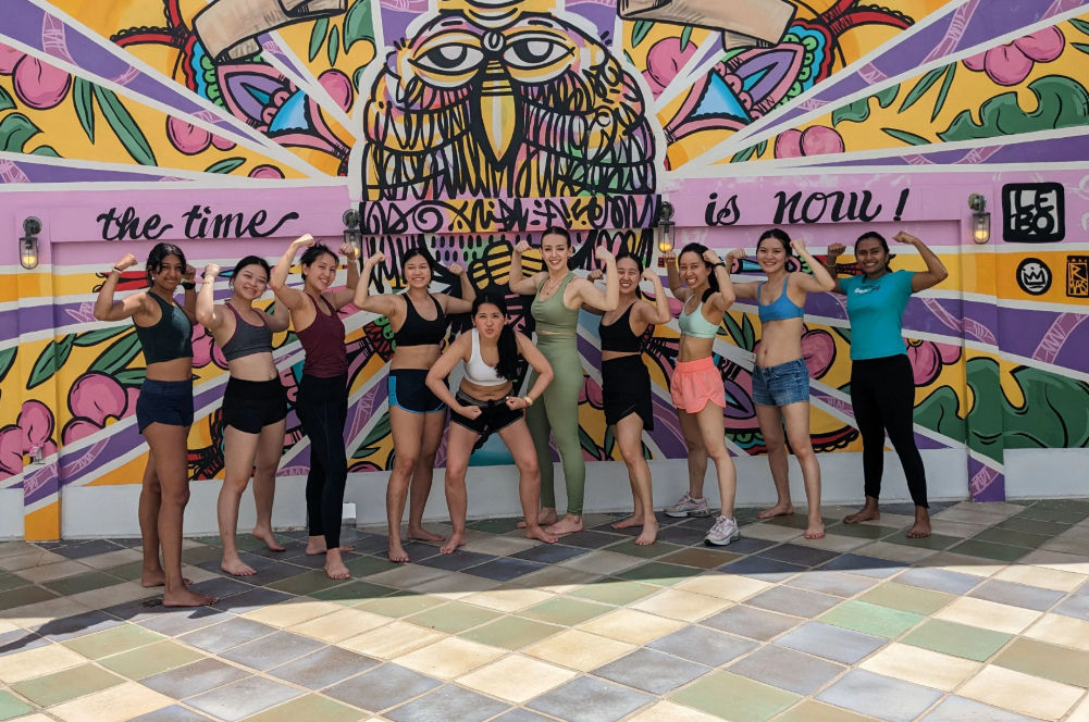 Energetic group of women in workout gear flexing and posing in front of a vibrant owl street mural that reads “the time is now,” standing on a tiled outdoor courtyard.