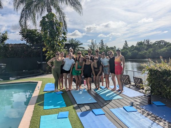 Group of women posing on blue yoga mats poolside by a waterfront canal under a palm tree on a sunny day — outdoor yoga / wellness gathering