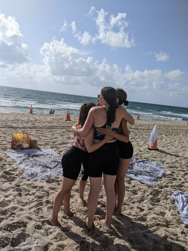 Four friends in swimsuits sharing a group hug on a sunny sandy beach with ocean waves, towels laid out and orange safety cones under a blue sky with scattered clouds.