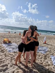 Four friends in a group hug on a sunny sandy beach, wearing swimsuits and shorts with towels nearby, ocean waves and blue sky with fluffy clouds in the background.