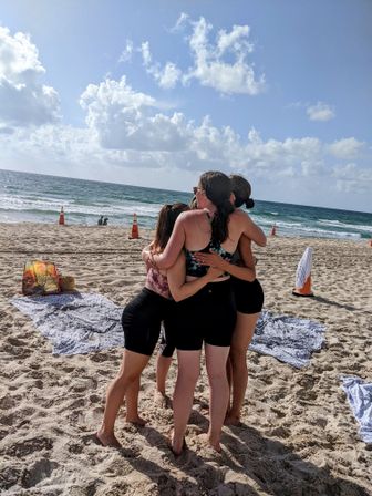 Four friends in a group hug on a sunny sandy beach, wearing swimsuits and shorts with towels nearby, ocean waves and blue sky with fluffy clouds in the background.