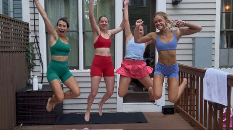 Four friends in colorful activewear jumping mid-air on a residential backyard wooden deck in front of a house door during an outdoor home workout
