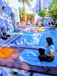 Sunny poolside yoga class in a tropical courtyard — group seated meditation on mats around a small pool with colorful inflatable floats, palm trees, and a black-and-white wall mural.