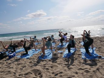 Group practicing yoga on a sandy beach on blue towels, balancing one-legged poses toward the sparkling ocean under a sunny sky