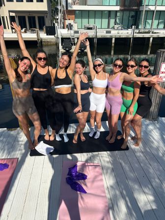 Eight women in colorful activewear smiling and posing with arms raised on a sunny waterfront dock by a residential canal, yoga mats on the deck and modern homes and boat lifts in the background.