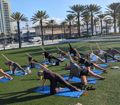 Group outdoor yoga class on blue mats on a sunny coastal park lawn, participants stretching in side poses among palm trees with nearby high-rise buildings and a promenade in the background.