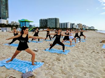 Energetic group beach yoga class doing warrior pose on blue-striped towels on a sandy shore with a colorful lifeguard hut and coastal high-rise skyline in the background.