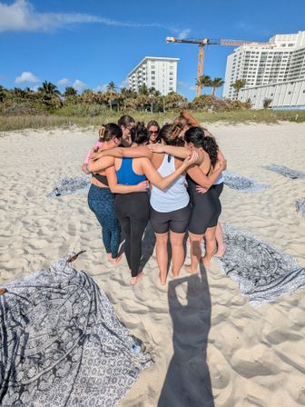 Group of women in activewear hugging in a circle on a sunny sandy beach with palm trees, patterned towels, and high-rise beachfront buildings and a crane in the background