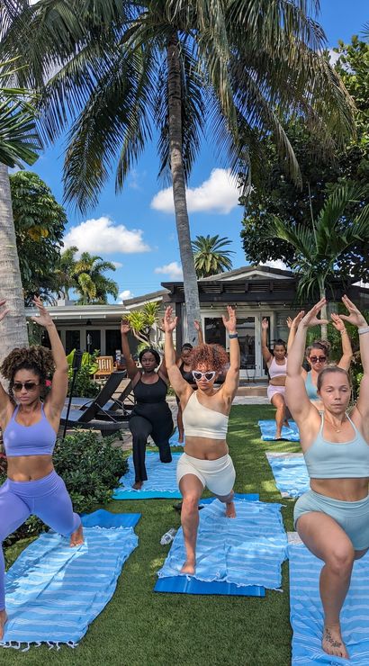 Outdoor yoga class of women doing lunges on blue striped mats in a sunny tropical backyard with palm trees and a pool