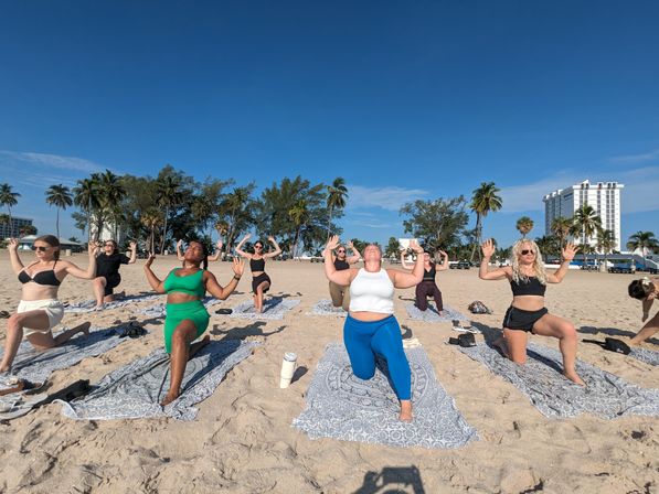 Beach yoga class on a sunny palm-lined waterfront — participants kneeling on patterned towels for outdoor fitness with palm trees and a high-rise building in the background.