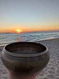 Bronze singing bowl held over a sandy beach at sunset, orange sun reflecting on calm ocean waves with cargo ships on the horizon — serene coastal meditation scene.