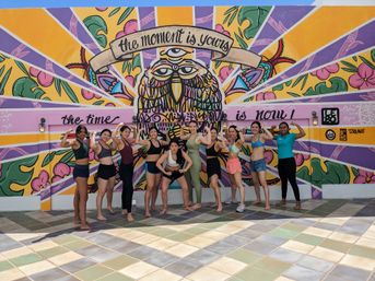Group of women in workout clothes flexing and posing on a sunny tiled patio in front of a colorful street-art mural featuring a sunburst owl and a banner reading “the moment is yours”.