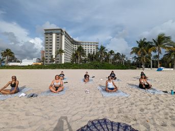 Group doing beach yoga on a sandy shore with palm trees and a large beachfront hotel under a blue, partly cloudy sky
