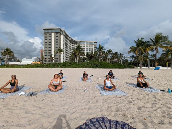 Group doing beach yoga on a sandy shore with palm trees and a large beachfront hotel under a blue, partly cloudy sky
