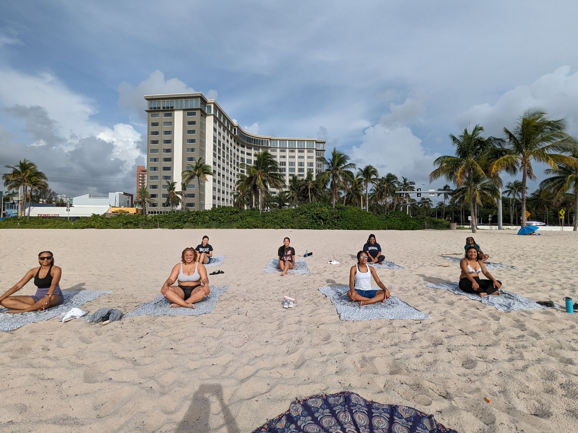 Beach yoga class on a sandy shore with palm trees and a curved multi-story beachfront hotel in the background under a partly cloudy sky — coastal Florida beach wellness scene.
