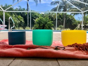 Bright outdoor sound‑healing setup with three crystal singing bowls (dark blue, teal, yellow) on a pink blanket beside a screened tropical pool deck with palm trees and small cymbals.