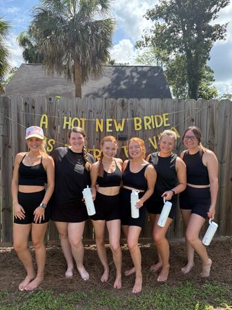 Six smiling women in matching black activewear standing barefoot in a sunny backyard, holding white water bottles in front of a wooden fence decorated with a gold "A HOT NEW BRIDE" banner and palm trees overhead.