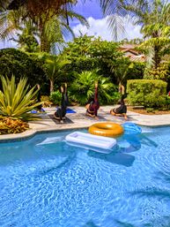 Poolside yoga: four people on mats in a sunny tropical backyard by a clear blue pool with palm trees and colorful inflatable floats.
