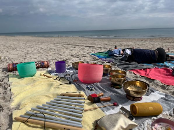 Vibrant crystal and brass singing bowls, chimes, mallets and blankets arranged on a sandy beach by the ocean for a relaxed sound-healing session under a cloudy sky.