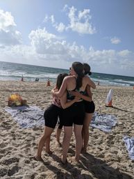 Four friends in swimsuits hugging in the sand on a sunny beach with towels, orange safety cones and ocean waves under a blue sky with fluffy clouds.