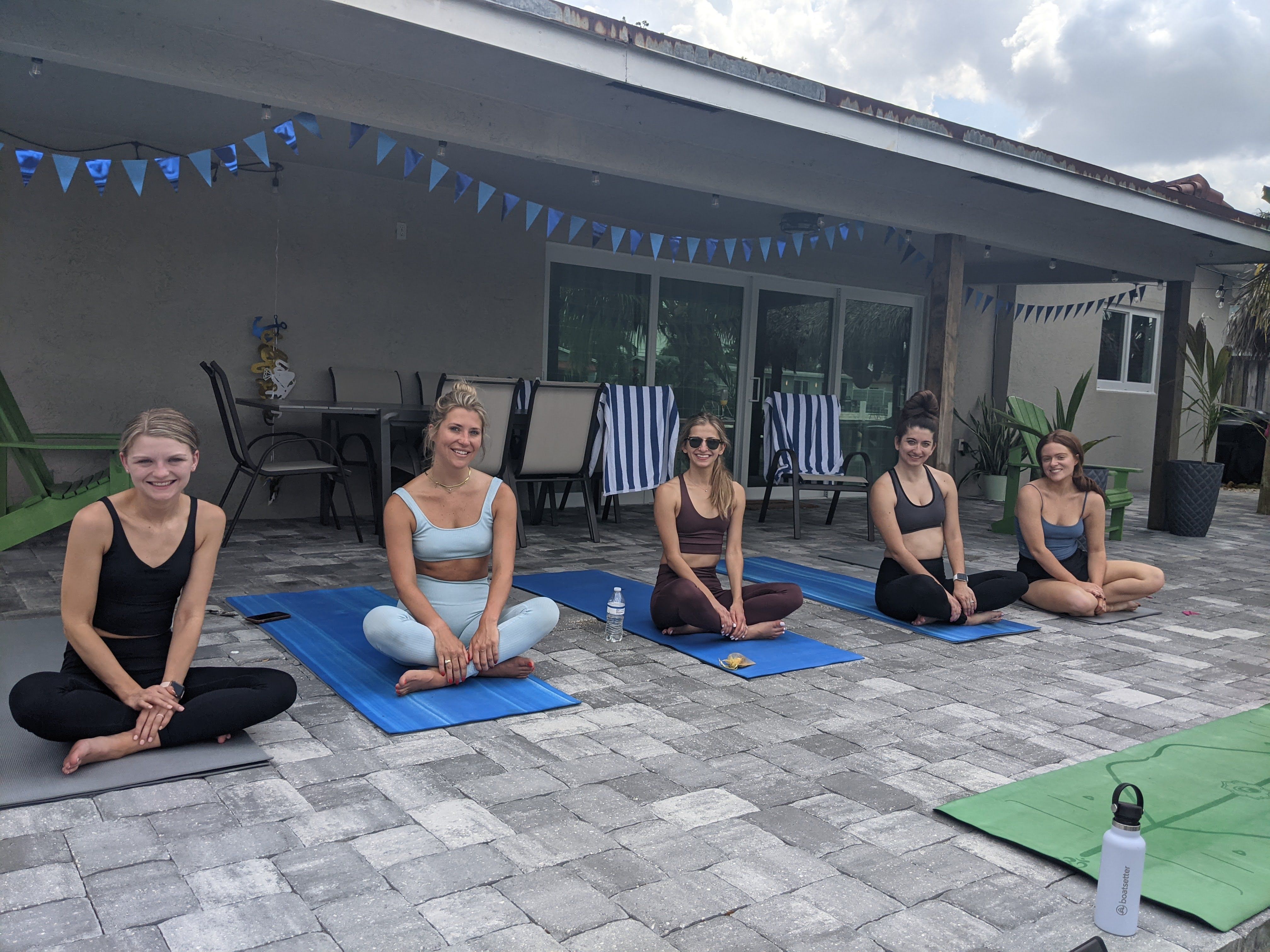 Five women seated cross-legged on yoga mats for an outdoor yoga session on a tiled backyard patio under a covered porch with blue pennant banners, striped lounge chairs and water bottle
