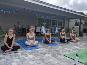 Five women seated cross-legged on yoga mats for an outdoor yoga session on a tiled backyard patio under a covered porch with blue pennant banners, striped lounge chairs and water bottle