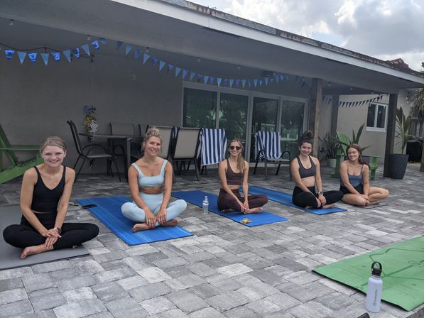 Five women seated cross-legged on yoga mats for an outdoor yoga session on a tiled backyard patio under a covered porch with blue pennant banners, striped lounge chairs and water bottle