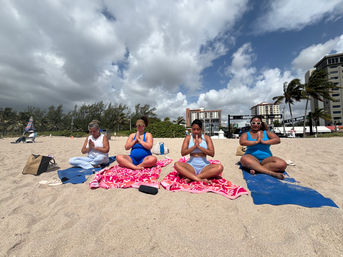 Group beach yoga: four women seated cross-legged on colorful towels meditating in prayer pose on a breezy oceanfront beach with palm trees and beachfront buildings under a partly cloudy sky.