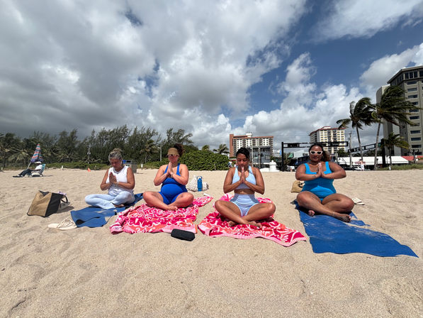 Group beach yoga: four women seated cross-legged on colorful towels meditating in prayer pose on a breezy oceanfront beach with palm trees and beachfront buildings under a partly cloudy sky.