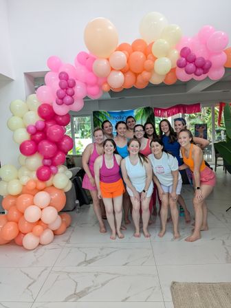 Smiling group of women in bright workout outfits posing barefoot under a large pastel pink, peach and cream balloon arch at an indoor tropical-themed party or fitness retreat.