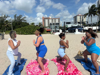 Four women in activewear doing partner yoga squats on colorful towels on a sunny palm-lined beach with coastal buildings and blue sky.