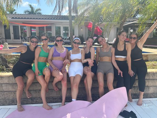 Eight women in colorful activewear posing on a wall by a sunny backyard pool with palm trees, enjoying a relaxed summer day.