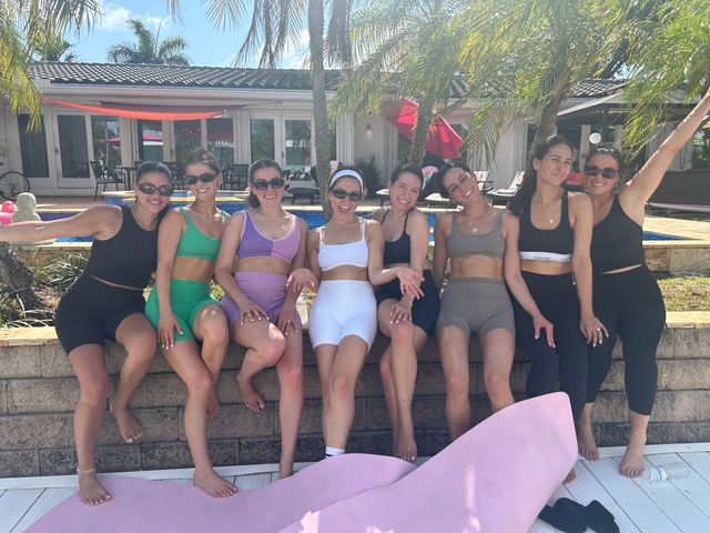 Eight women in colorful activewear posing on a wall by a sunny backyard pool with palm trees, enjoying a relaxed summer day.