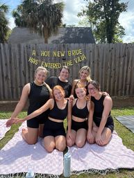 Six smiling women in black activewear kneeling on pink check blankets in a sunny backyard with palm trees, under a gold banner that reads A HOT NEW BRIDE HAS ENTERED THE VILLA — playful outdoor bachelorette party scene