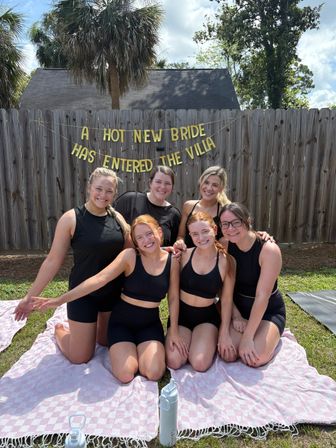 Six smiling women in black activewear kneeling on pink check blankets in a sunny backyard with palm trees, under a gold banner that reads A HOT NEW BRIDE HAS ENTERED THE VILLA — playful outdoor bachelorette party scene