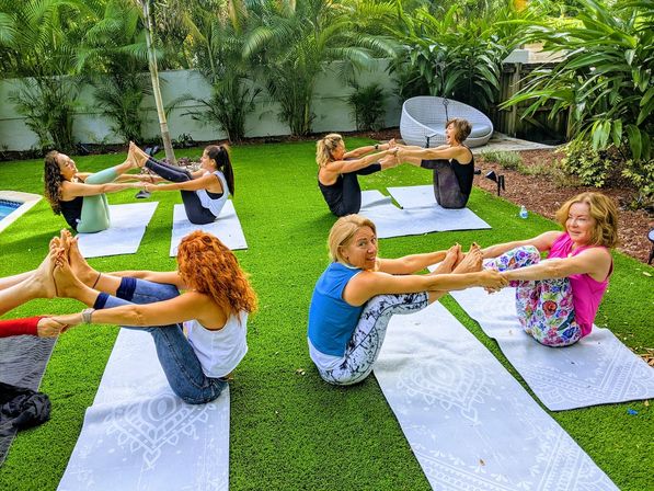 Smiling group of women doing partner boat-pose on yoga mats on green turf in a lush tropical backyard garden, outdoor group yoga session
