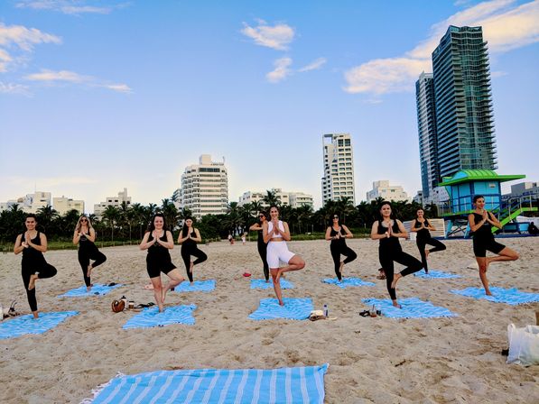 Beach yoga class on Miami Beach: people in tree pose on blue striped towels on sandy shoreline with palm trees, beachfront high-rise skyline and a colorful lifeguard tower under a clear sky.