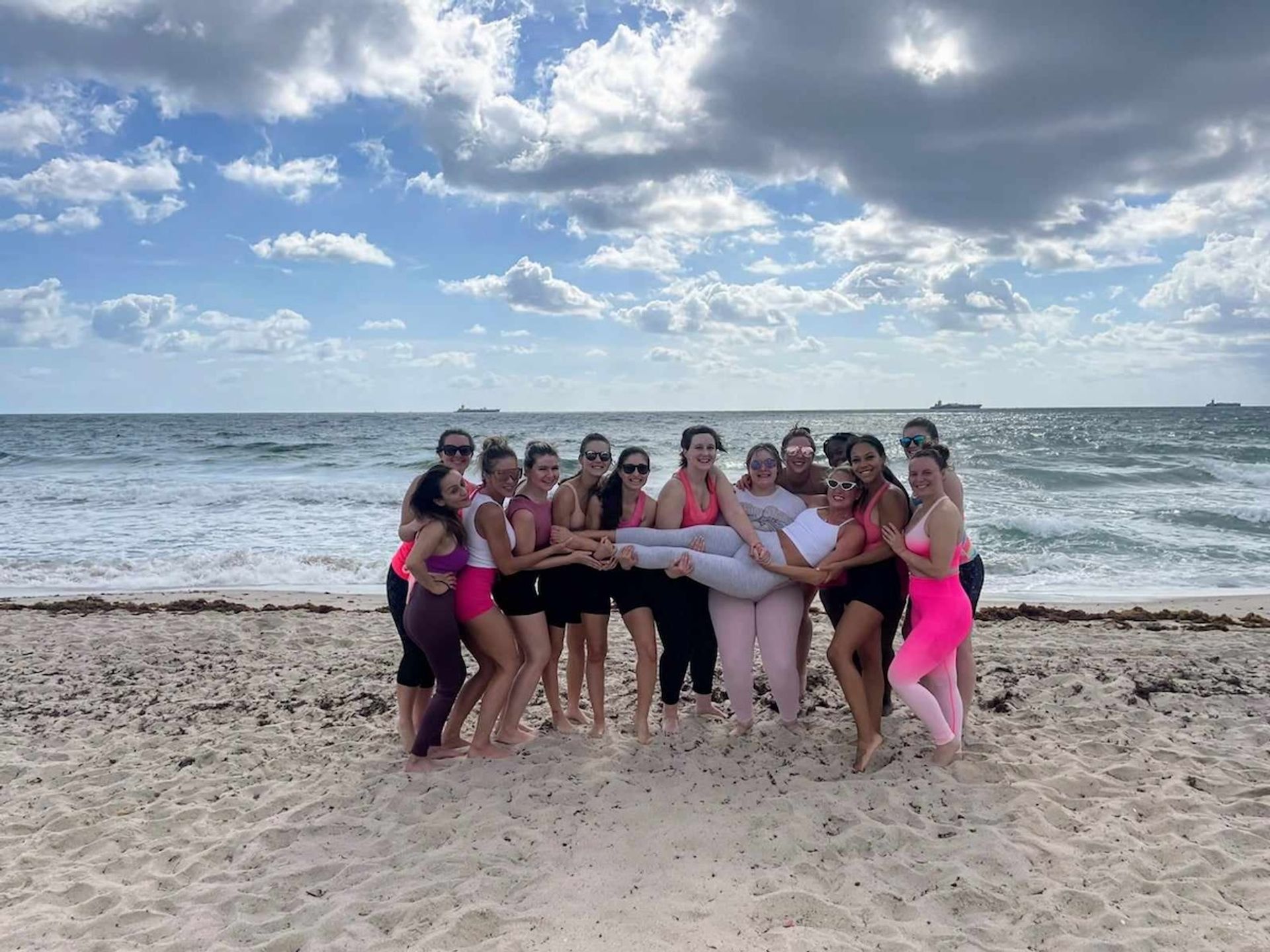 Group of friends in bright activewear on a sandy ocean beach under a dramatic cloudy sky, smiling and posing while lifting one friend horizontally above the sand.