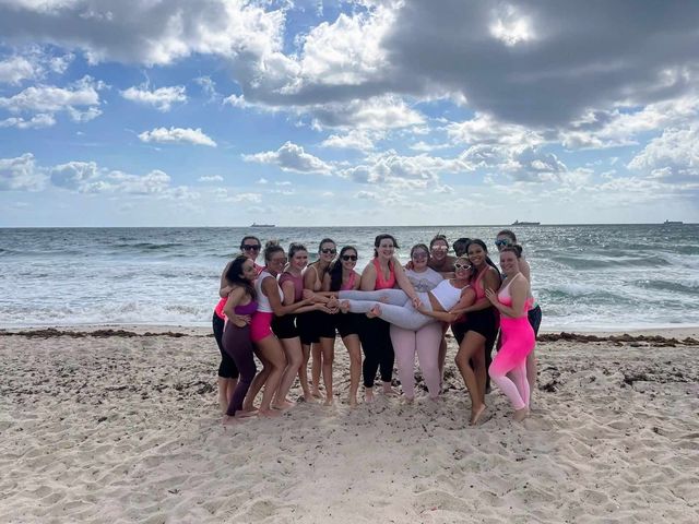 Group of friends in bright activewear on a sandy ocean beach under a dramatic cloudy sky, smiling and posing while lifting one friend horizontally above the sand.
