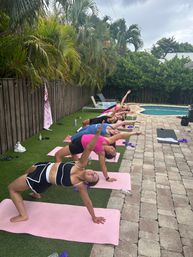 Smiling group fitness participants doing a poolside outdoor yoga stretch on pink mats, performing side-bridge poses with arms raised beside a backyard swimming pool and palm trees.