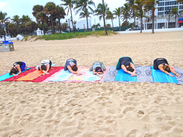 Six people in child's pose on colorful yoga mats during a group beach yoga session on a sandy shore with palm trees and oceanfront buildings in the background.