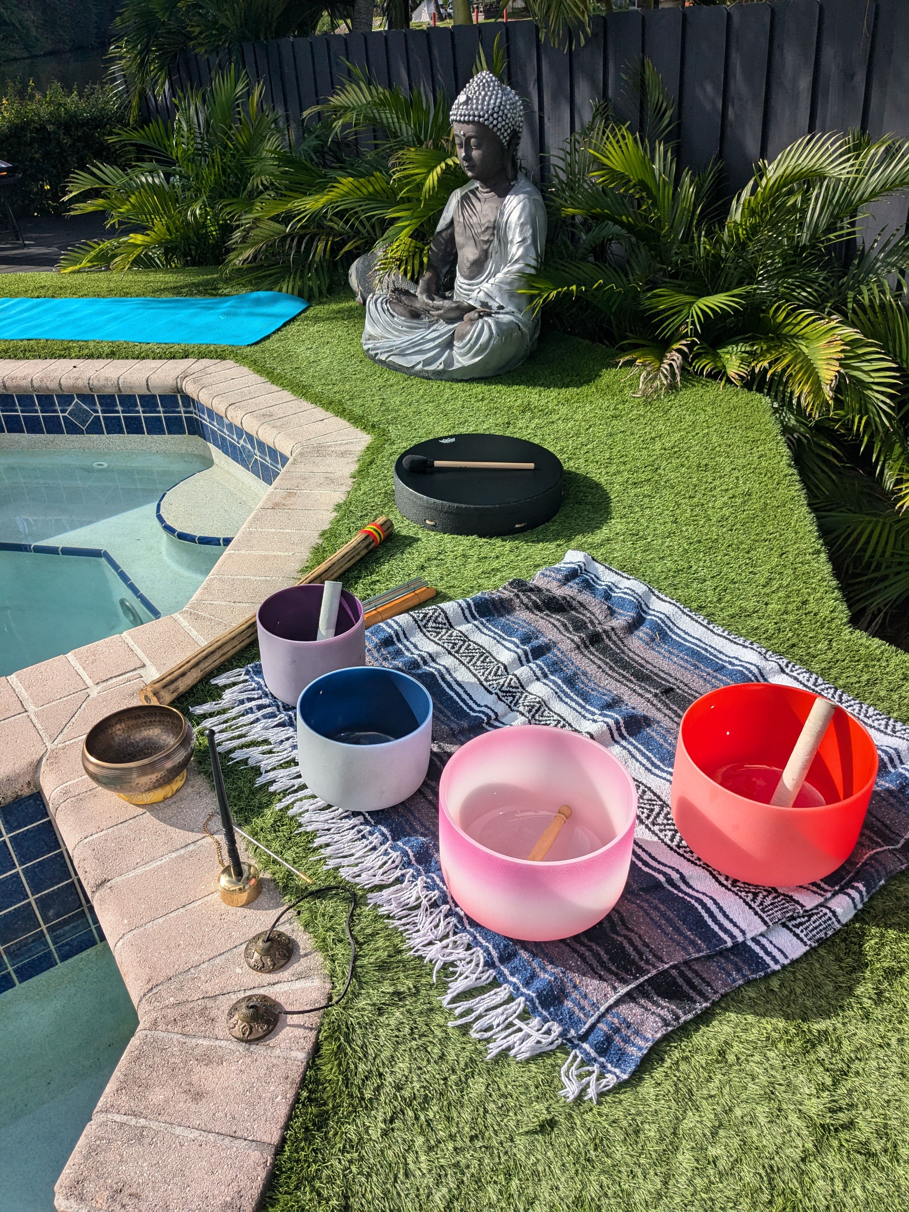 Poolside backyard meditation setup with a seated Buddha statue, colorful crystal singing bowls and mallets on a patterned blanket, Tibetan singing bowl and tingsha cymbals on artificial grass beside a blue yoga mat and tropical palms.