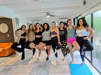 Eight women smiling and striking tree pose in a bright indoor yoga class, lined up on mats by large windows in a sunlit studio.
