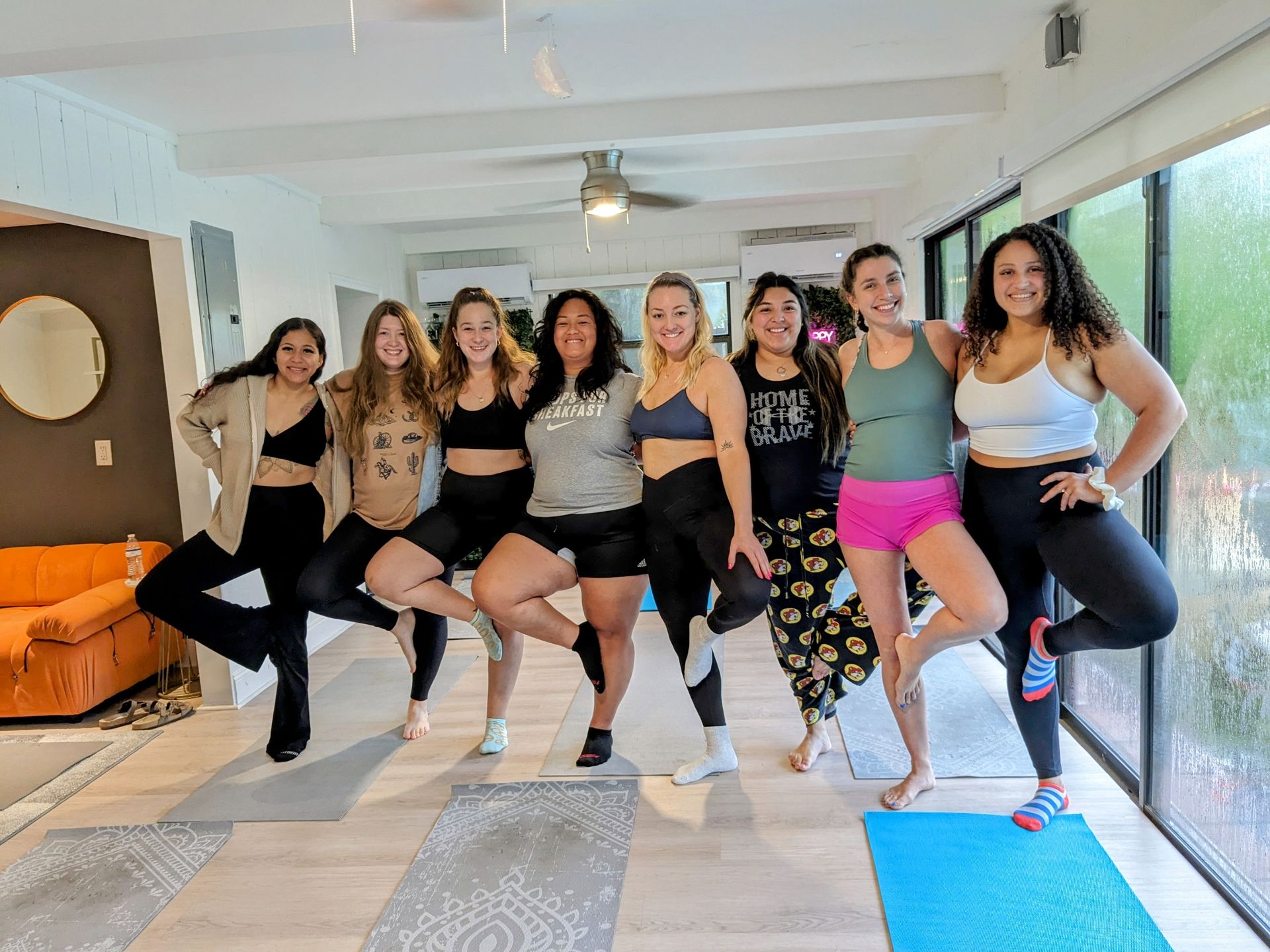 Eight smiling women in athletic wear holding tree pose together in a bright indoor yoga studio with floor-to-ceiling windows, yoga mats, and light wood floors — group fitness/yoga session.