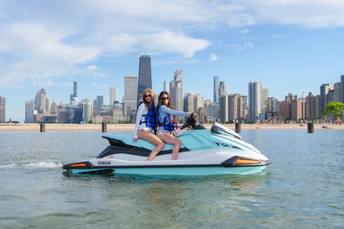 Two friends in life jackets on a turquoise jet ski on Lake Michigan with Chicago skyline and sandy beach in the background on a sunny summer day