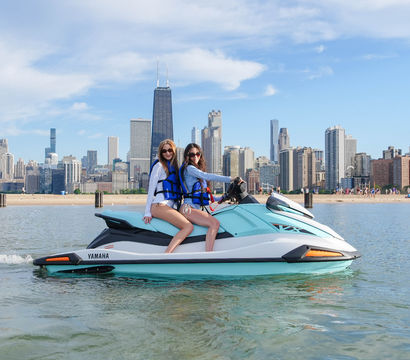 Two friends in life jackets on a turquoise jet ski on Lake Michigan with Chicago skyline and sandy beach in the background on a sunny summer day
