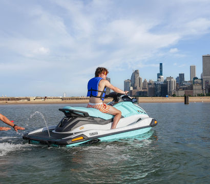 Rider in a blue life vest on a turquoise jet ski being pushed off the shallow water by a helper, sandy lakefront and Chicago skyline along Lake Michigan with tall skyscrapers under a partly cloudy sky
