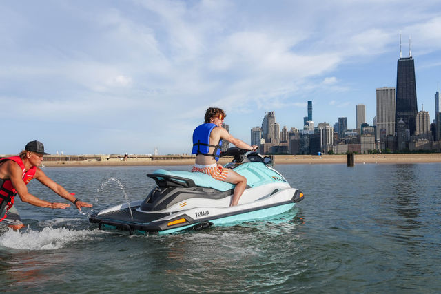 Rider in a blue life vest on a turquoise jet ski being pushed off the shallow water by a helper, sandy lakefront and Chicago skyline along Lake Michigan with tall skyscrapers under a partly cloudy sky
